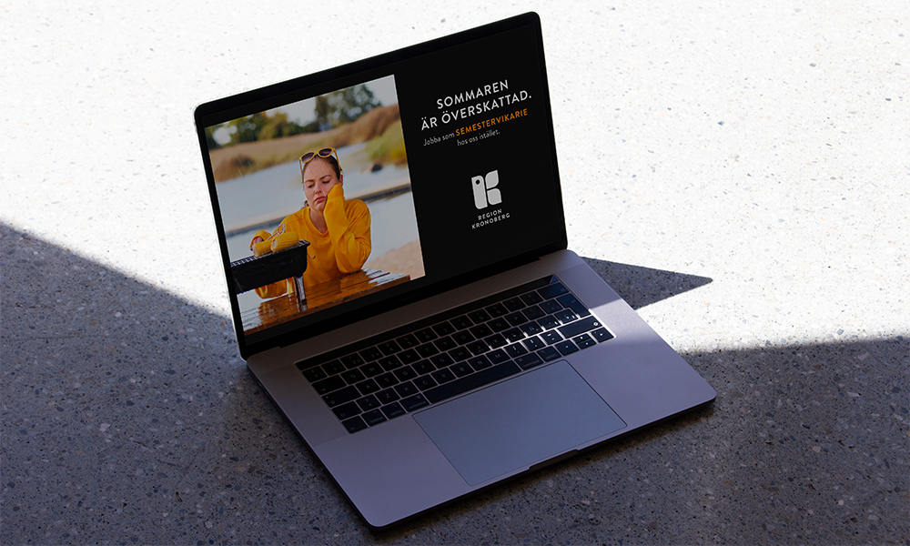 En MacBook Pro står på ett betonggolv i halvskugga. På skärmen visas en kampanjbild från Region Kronoberg: En ung kvinna i gul tröja ser uttråkad ut vid en strand med en liten grill framför sig. Bredvid står texten: "Sommaren är överskattad. Jobba som semestervikarie hos oss istället." Nedanför visas Region Kronobergs logotyp.