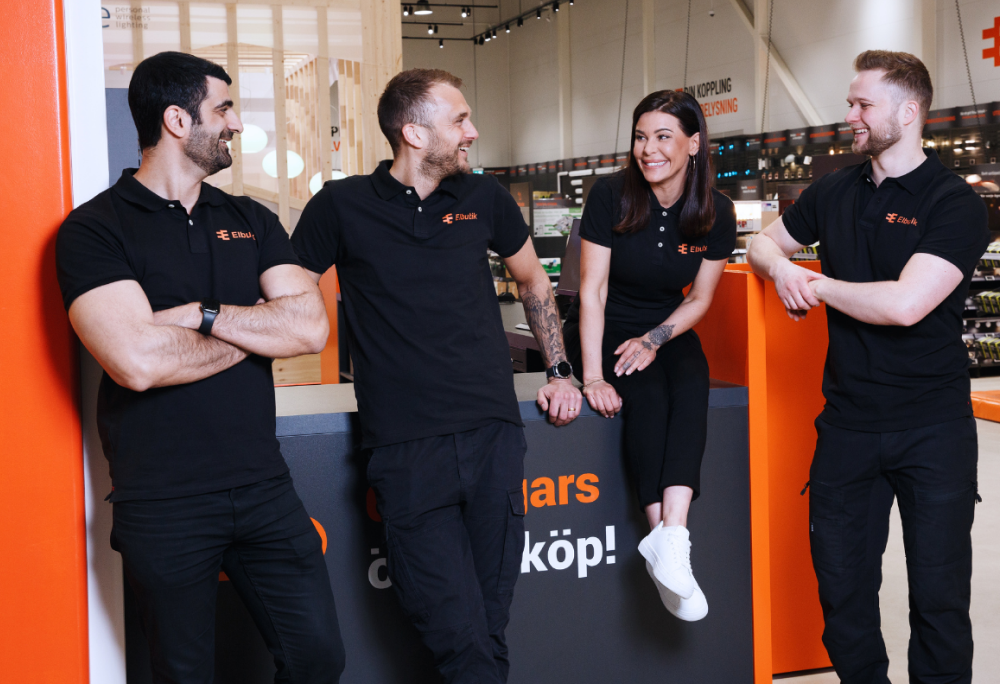 Fyra personer i svarta Elbutik-tröjor står och pratar glatt i en butiksmiljö med orange och vita detaljer. Till vänster syns Elbutiks logotyp mot orange bakgrund.