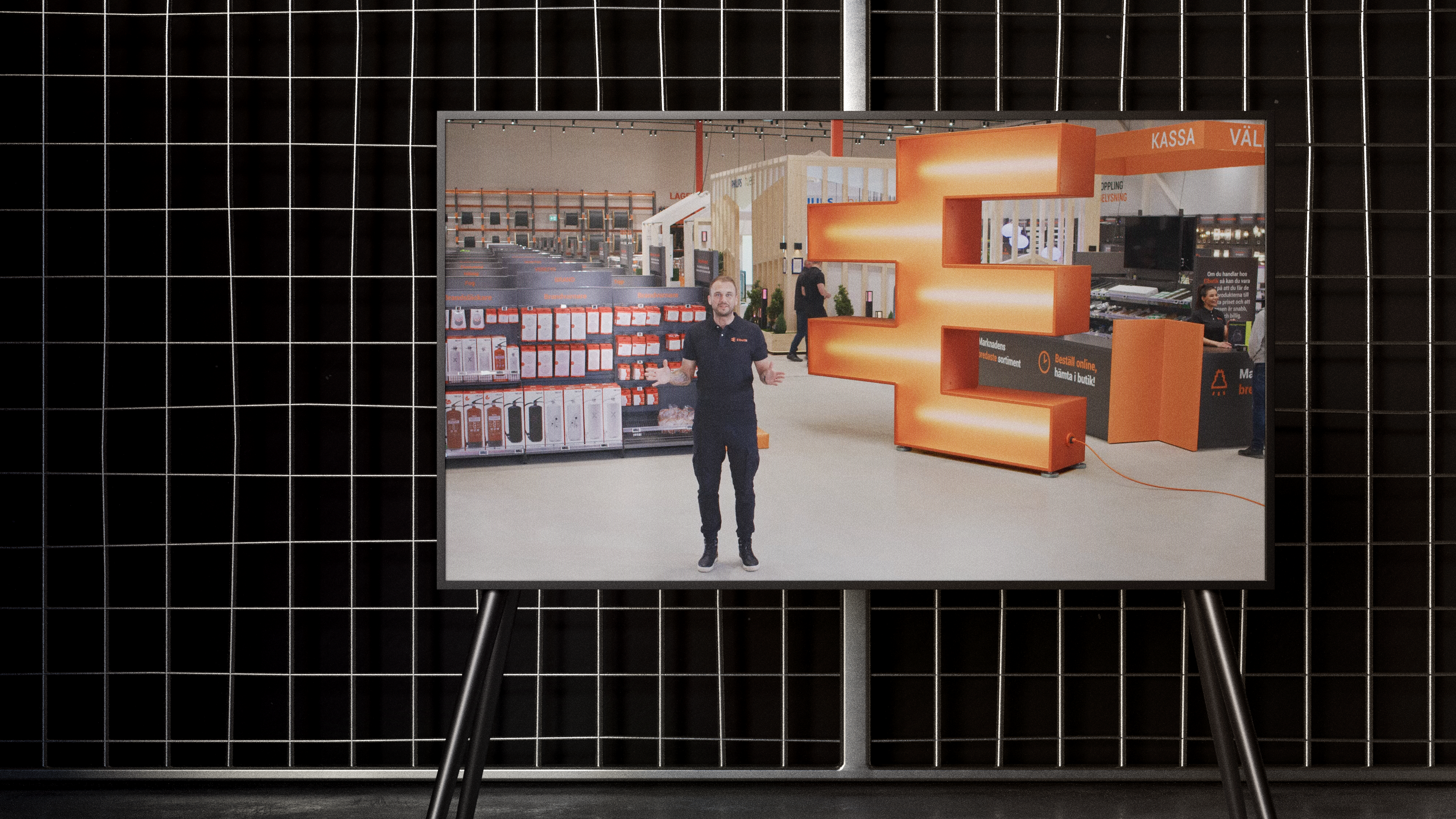 En tv-skärm visar en person som står i Elbutiks butik framför en stor upplyst E-symbol i orange. Bakom syns hyllor med produkter och butikens kassaområde.